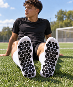 A man sitting on a green soccer field wearing white socks with black circular studs on the soles, looking off-camera. The background shows a soccer field, goal, trees, and blue sky.